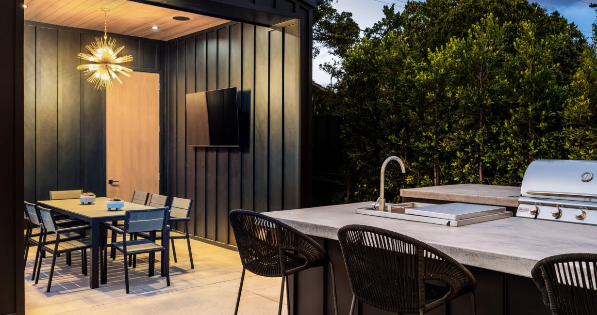 Modern outdoor kitchen and dining area with sleek furniture, grill, and stylish lighting, surrounded by lush greenery.